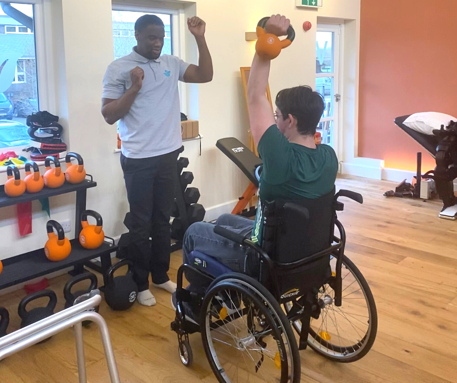 Jack in a personal training session with ash. He is sat in his wheel chair whilst holding a kettle bell above his head with his left arm. Ash is stood in front of him.