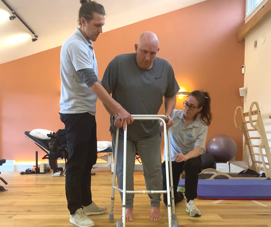 Stroke Survivor Jamie is pictured using his zimmer frame whilst physios tom and bex support him either side.