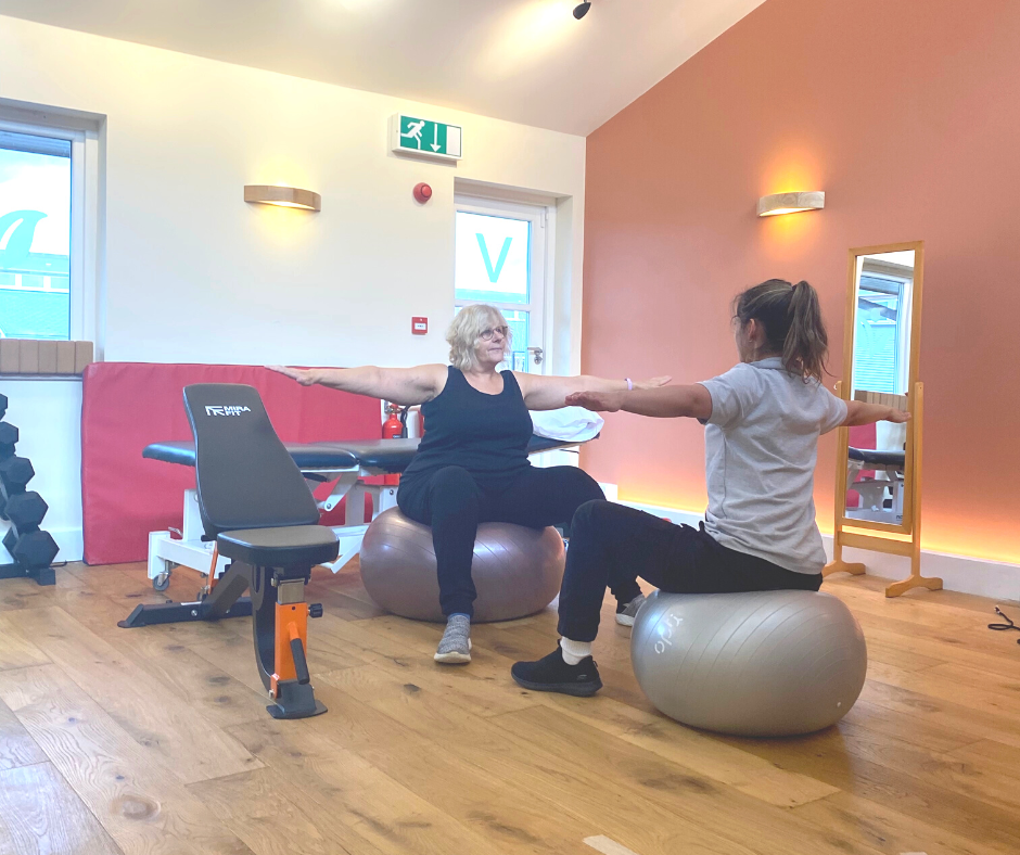 Woman sits on exercise ball whilst in personal training session her arms are stretched out to either side there is a physio in front of her doing the same movemnet
