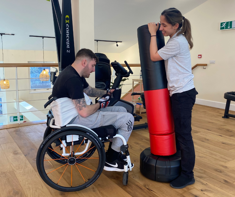 Shaun is boxing in his personal training session. He is sat in his wheel chair whilst physio maria holds up a punching bag.