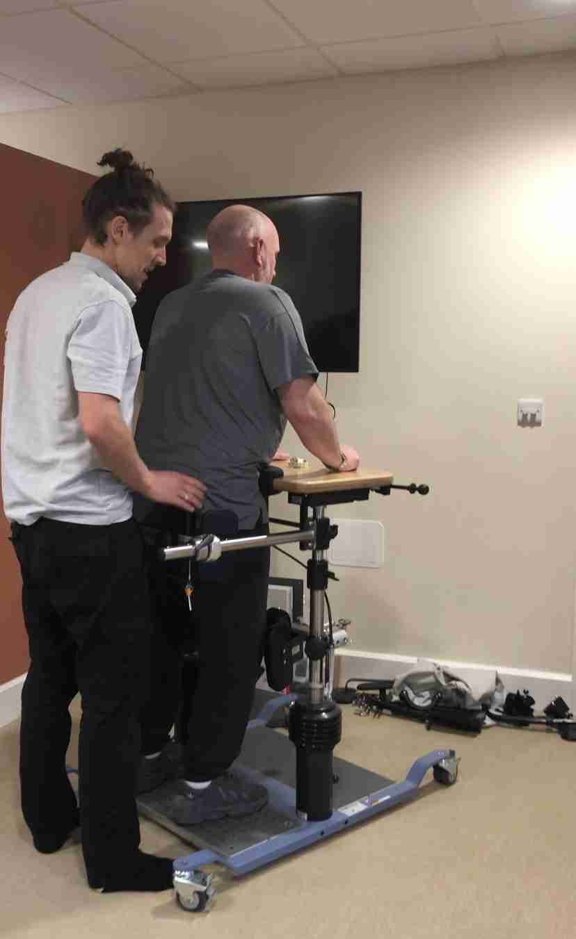jamie is stood in rehab tech device the balo, whilst he works on his balance and core. physio tom is stood behind him