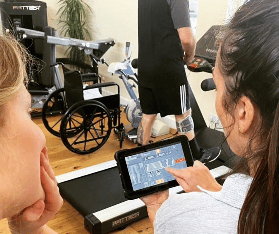 photo taken over the shoulder of physios bex and sophie who are looking at data from the bioness go on a small tablet. In the background we can see the bottom half of someone using the treadmill with the bioness attached to their right leg to help with Food Drop.