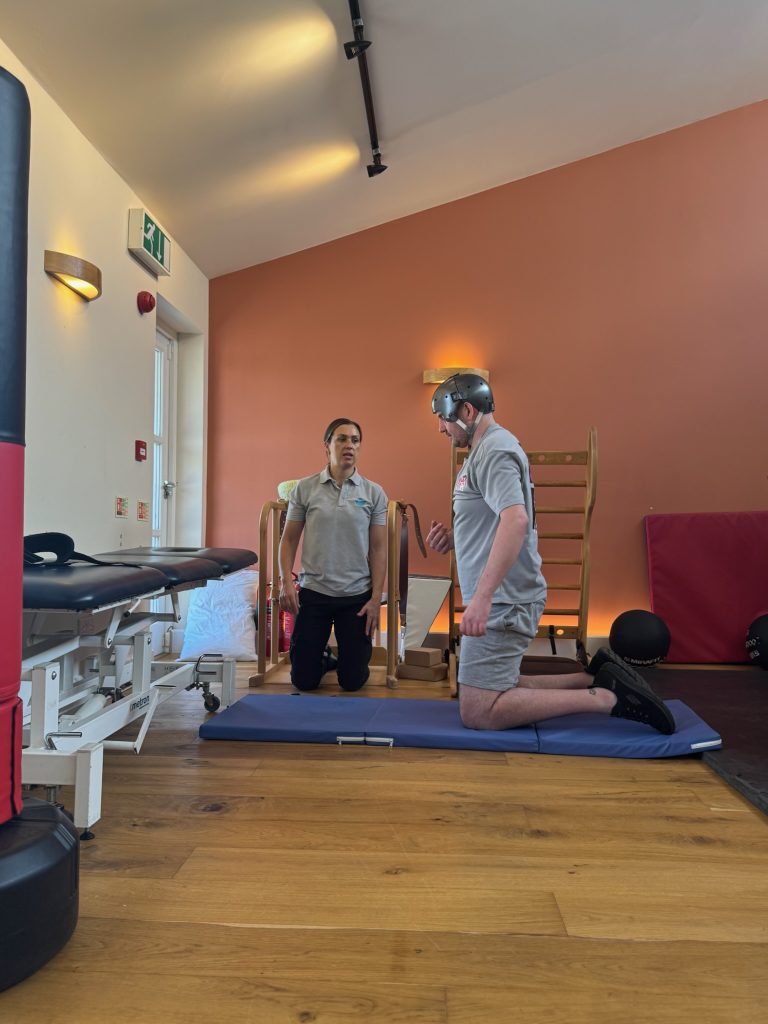 Oli is knelt up in his physio session next to physiotherapist Charlotte. he is wearing his helmet to protect his head.