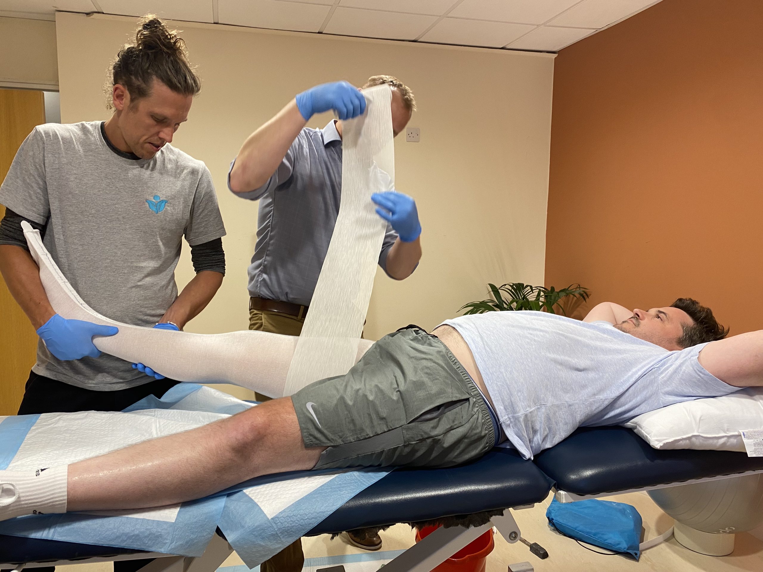 sid is lying on his back whilst physio tom holds his right leg and orthotics specialist wraps his leg in a cast