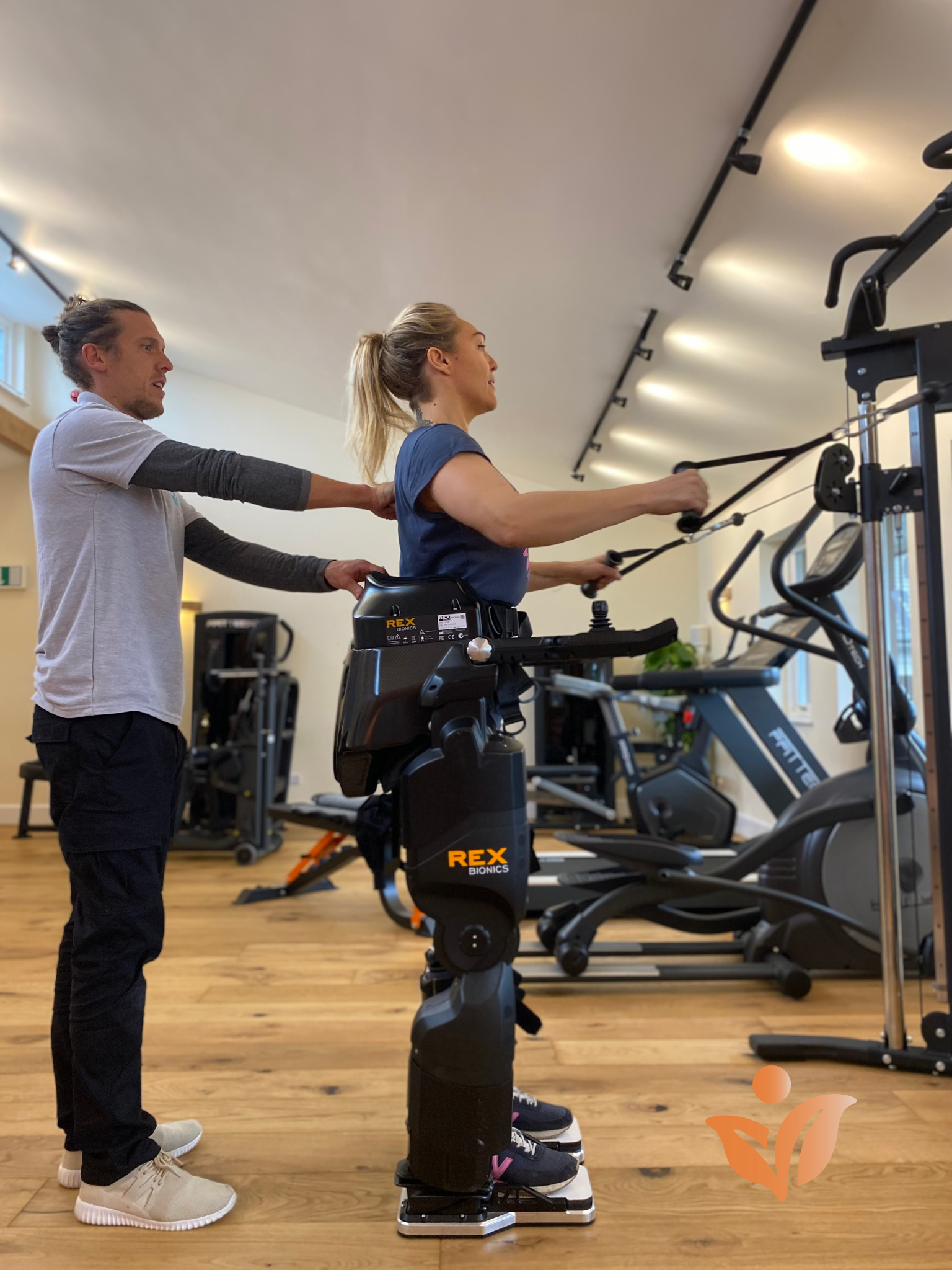 Sophie Morgan stands supported in the Rex exoskeleton whilst using the pulley weights. Physio Tom is Stood behind her.