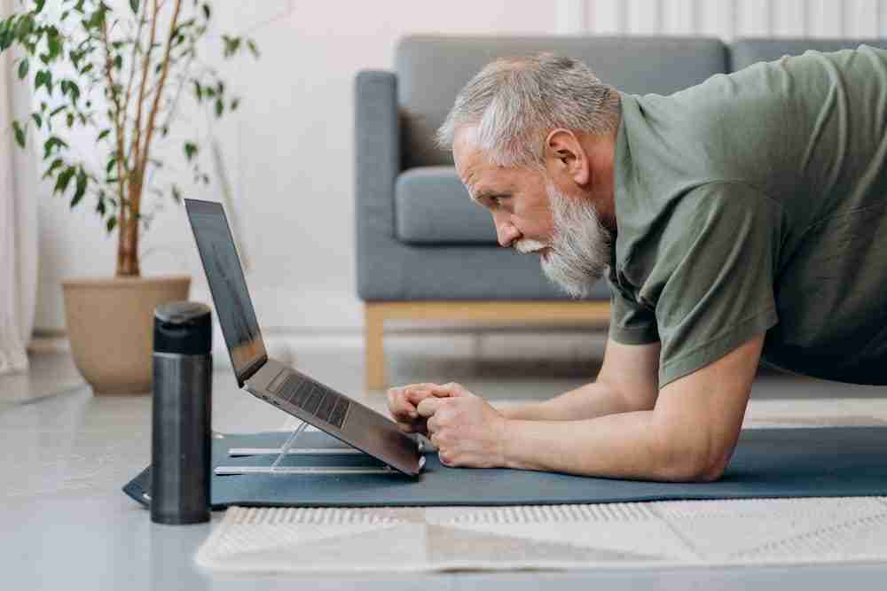 man crouched down on a yoga mat whilst looking at a laptop
