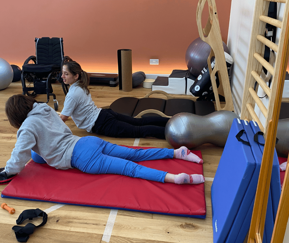Asha is on her belly on the floor in upwards dog with Pilates instructor Maria beside her.
