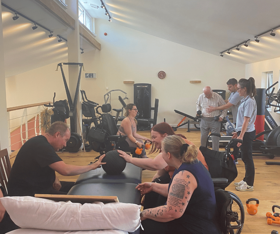 adaptive hiit class in full swing. 3 members sit passing a medicine ball to one another and in the background you can see to other gym members using the free weights.