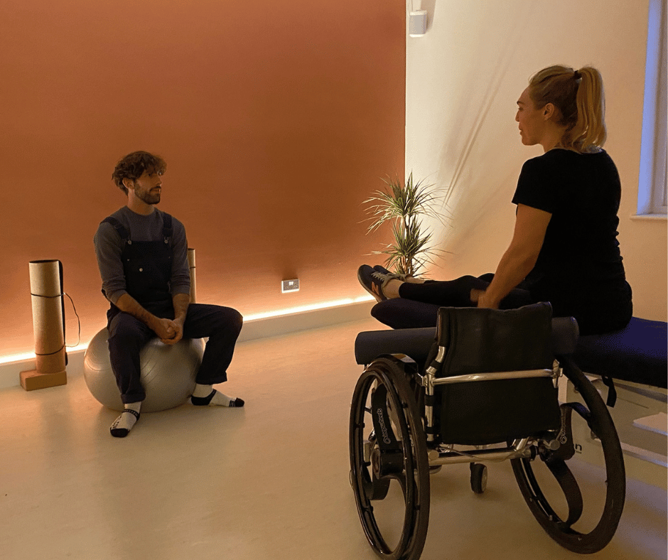 John sits in front of sophie morgan in a dimly light room. sophie is sat on a bed facing john with her legs stretched out in front of her and her wheel chair beside her.