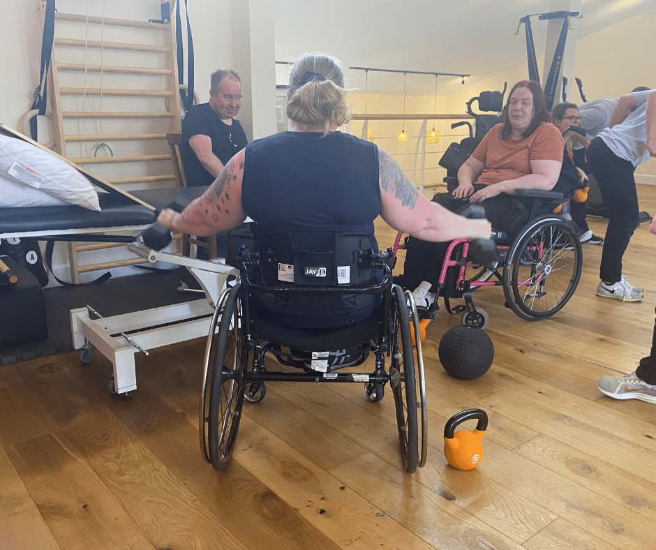 sarah sits in her wheelchair whilst using a free weight in each arm. she has her back to the camera