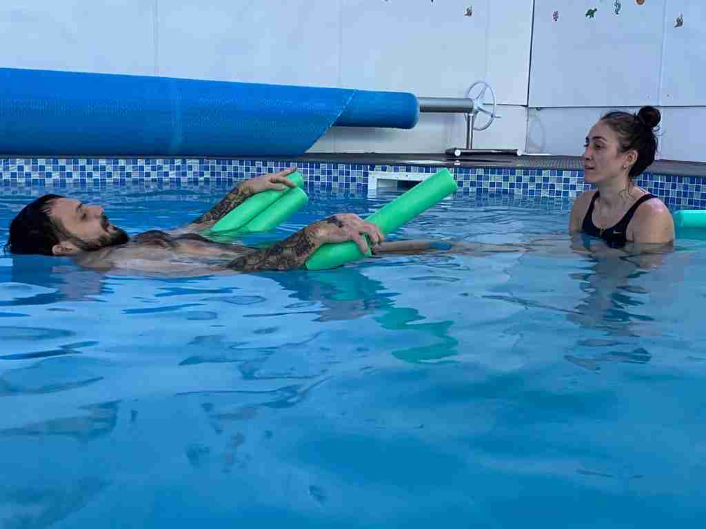 Elliot floats on his back whilst supported by a floatation device with physio bex positioned at his feet