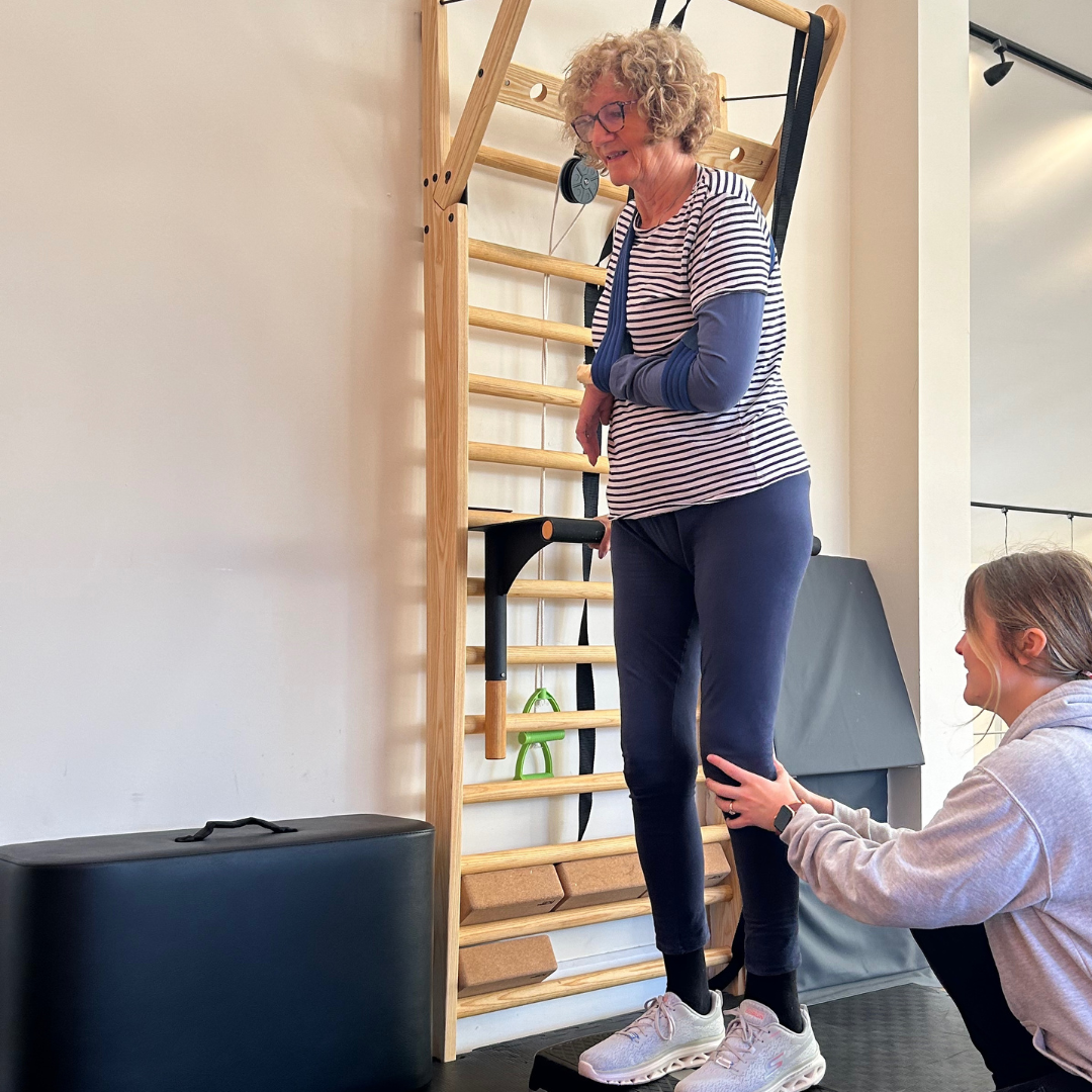 Christine is stood on an exercise step whilst holding onto a wooden wall bar for support. Her left arm is an a blue sling. Physio sophie is crouched by her side for support.