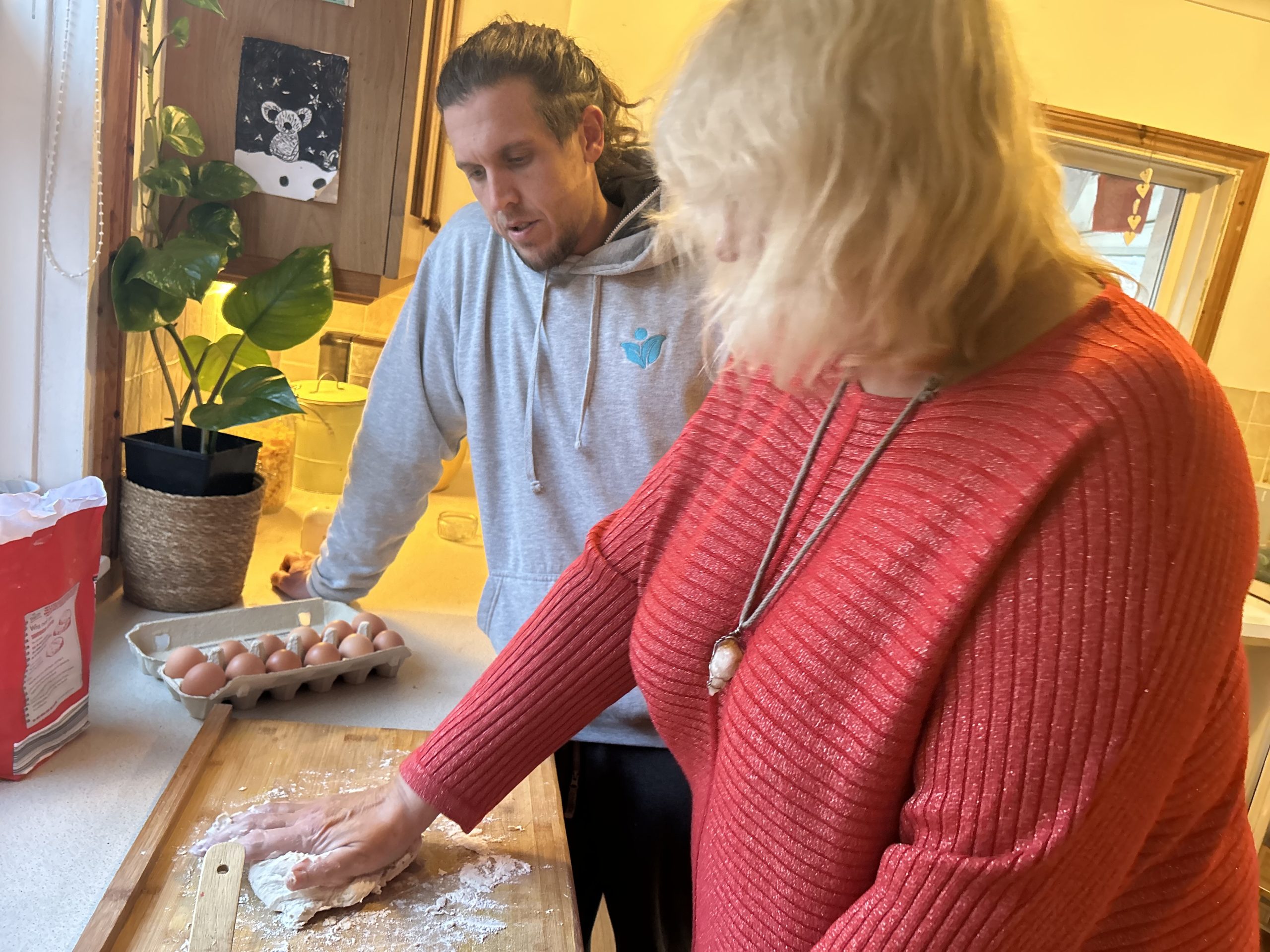 Julie is stood in her kitchen kneading dough with her left hand as part of her rehabilitation exercises at home. Physiotherapist tom is stood beside her