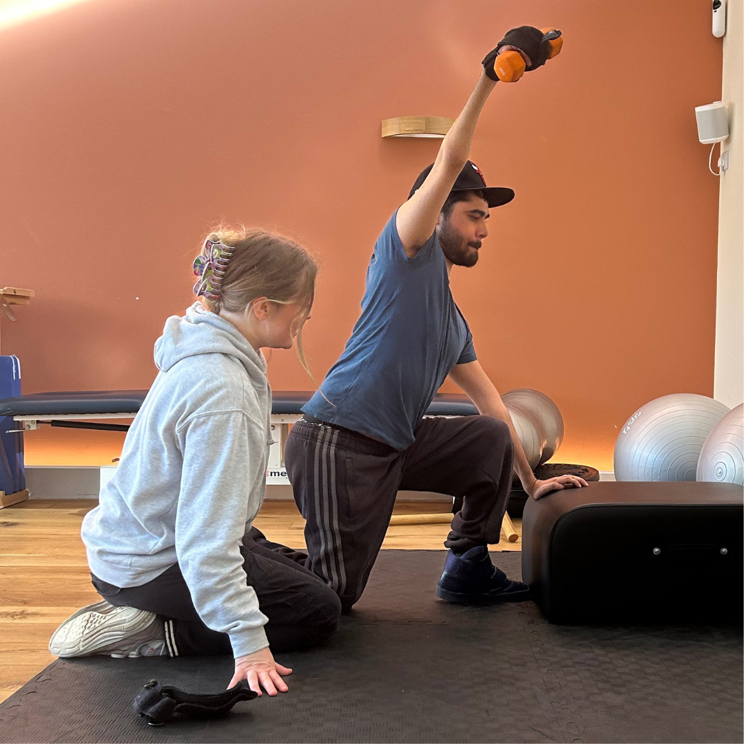 Ryan is knelt on one knee whilst raising a kettle bell with his right arm above his head. physio sophie is knelt beside him