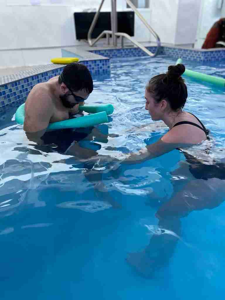 Elliot is in the hydrotherapy pool wearing googles whilst supported by a floatation noodle and Physiotherapist Bex