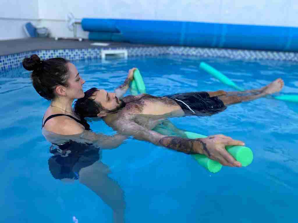 Elliot is floating on his back in the hydrotherapy pool whilst supported by a floatation device and physio Bex