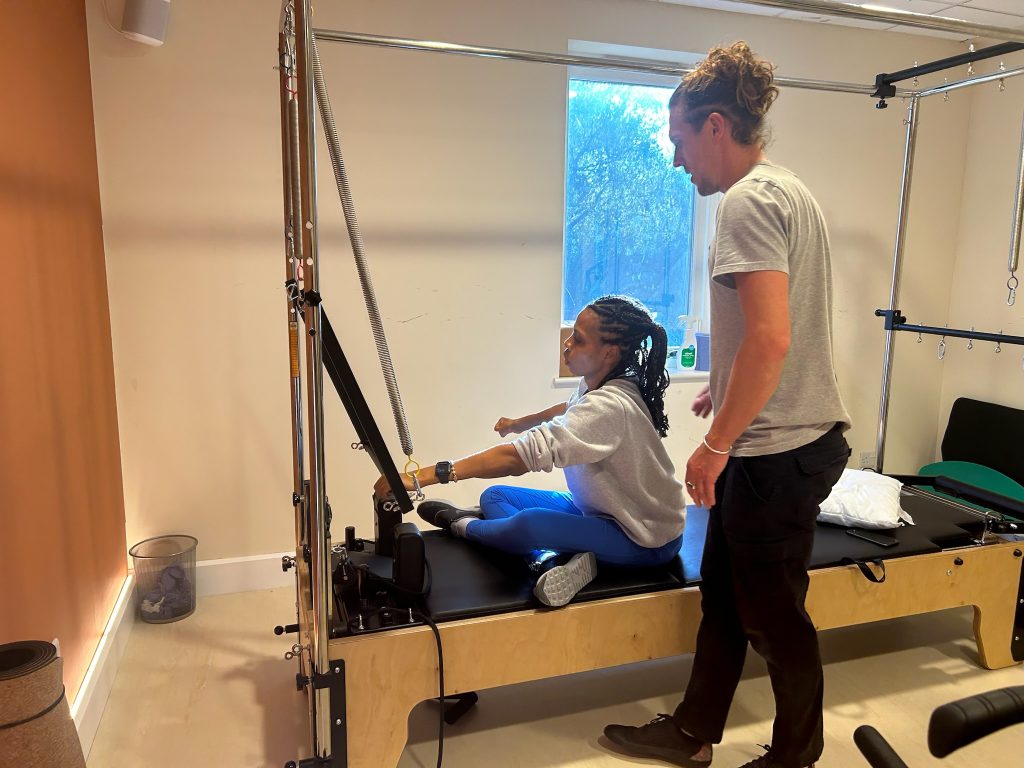 a black woman is sat cross legged on the pilates table whilst using the front bar todo a foward stretch.
